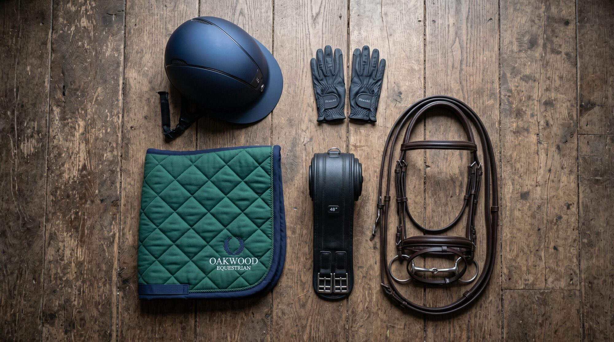 Overhead flat lay of essential horse tack — a riding helmet, gloves, a green saddle pad, a girth, and a bridle — arranged on a wooden barn floor